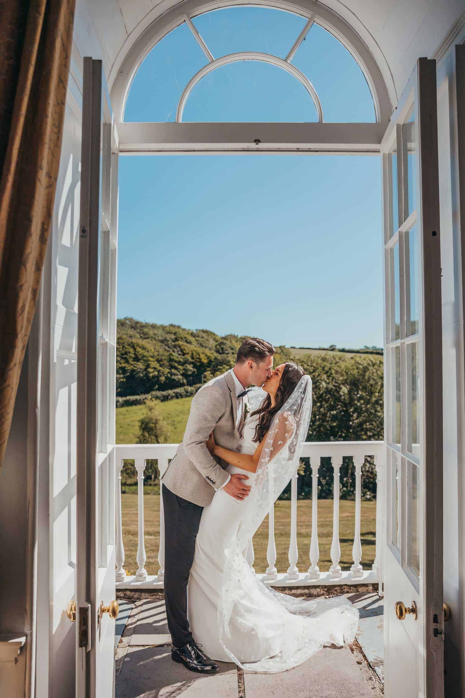 Bride and groom kissing at a window with scenic countryside view.