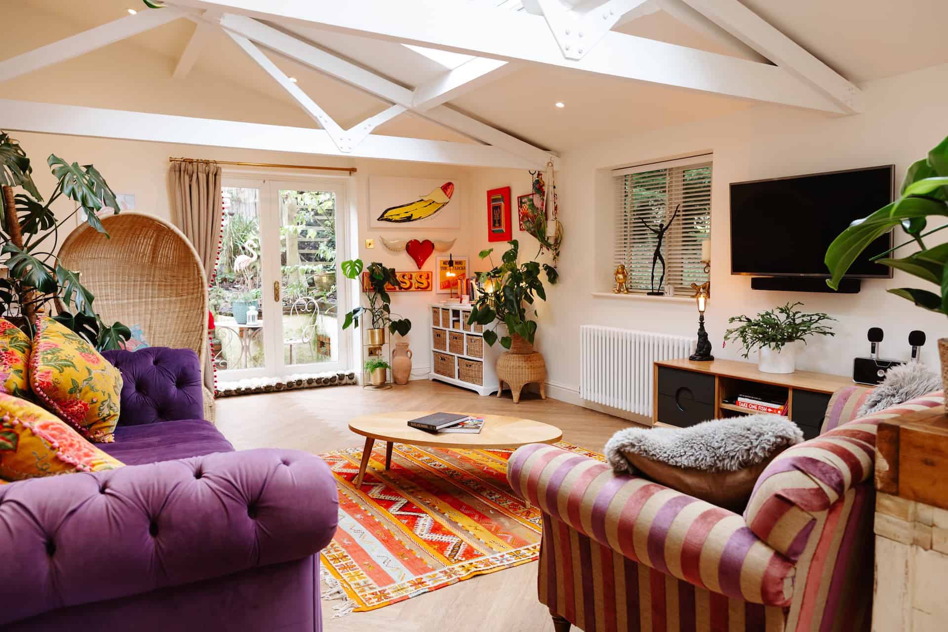 Bright, colourful living room with purple and striped sofas, plants, and wall art.