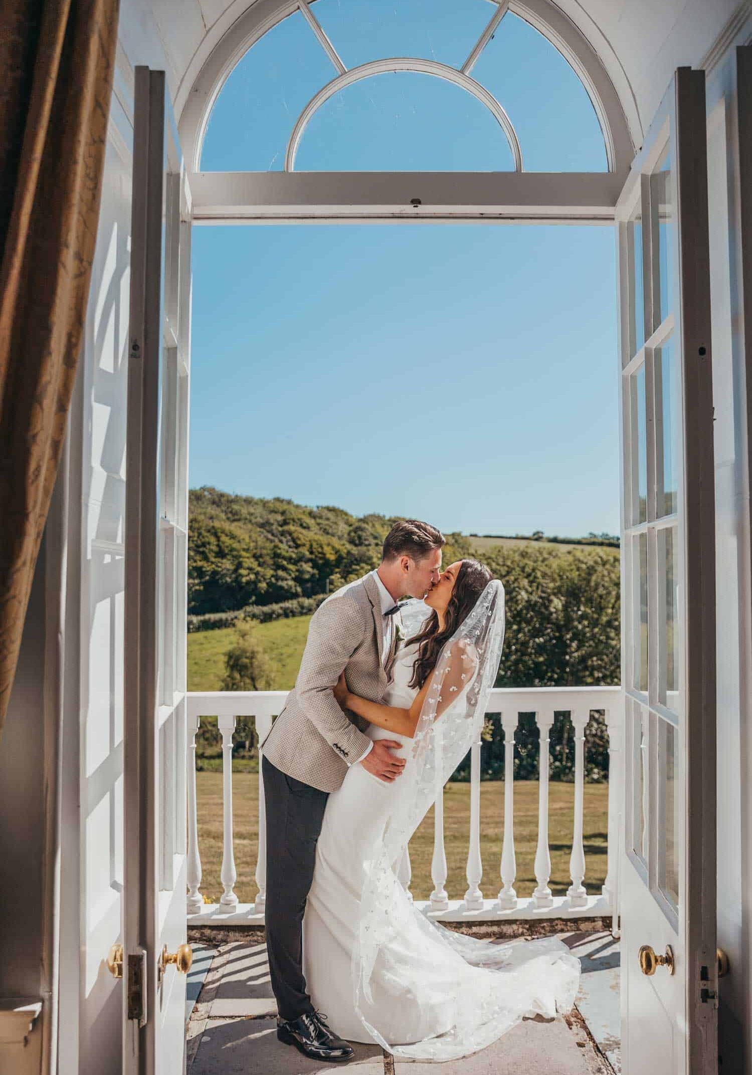 Bride and groom kissing at a window with scenic countryside view.