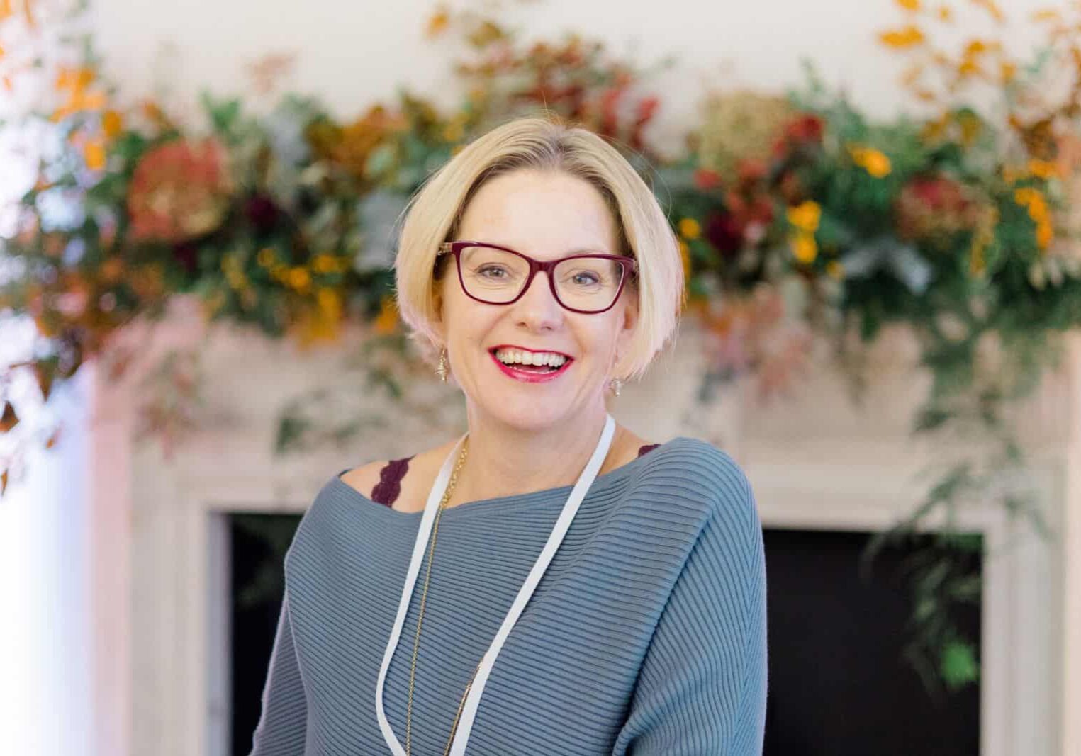 Woman wearing blue jumper standing in front of flowers smiling at camera | Kelly Chandler Success Coach